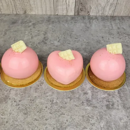 Trzy pastelowo różowe monoporcje, dwie okrągłe i jedna w kształcie serca, każda z kawałkiem białej czekolady.