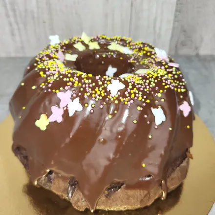 Babka w czekoladowej polewie, obsypana kolorowymi perełkami i cukrowymi motylkami, na złotej podkładce.