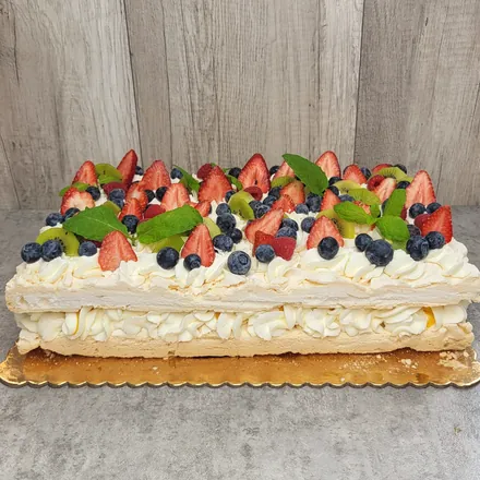 Prostokątny tort bezowy z bitą śmietaną, truskawkami, borówkami, kiwi, malinami i listkami mięty na złotej tacy.