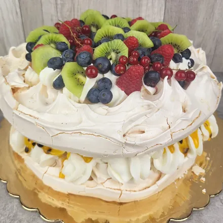 Okrągły tort bezowy z kiwi, malinami, porzeczkami i borówkami na wierzchu oraz marakują między blatami.