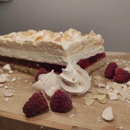 Kawałek ciasta z warstwą malin, kremem i bezą z płatkami migdałów, obok beziki i świeże maliny.