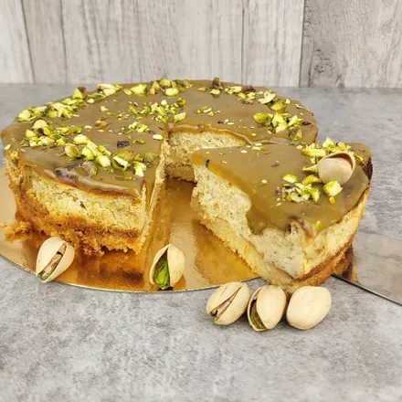 Sernik z pistacjową polewą i siekanymi pistacjami na złotej podkładce, z wyciętym kawałkiem.