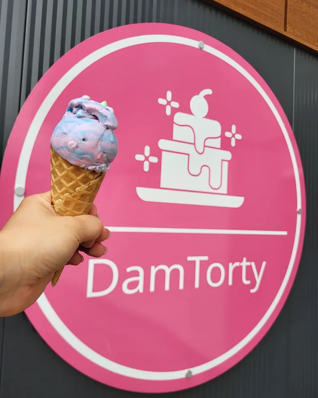 Lody w wafelku trzymane w dłoni przed różowym szyldem DamTorty